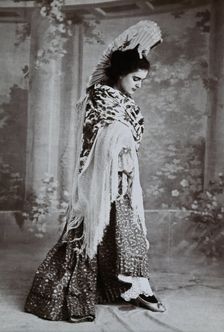 A standing woman, posed with an upraised fan and pointed toe, wearing a fringed shawl, in..., c1900. Creator: Unknown