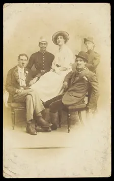 Amateur actors sitting down, surrounding a man in drag seated in the centre, [1910-1919?]. Creator: Unknown