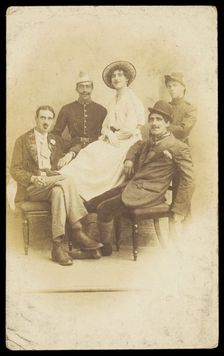 Amateur actors sitting down, surrounding a man in drag seated in the centre, [1910-1919?]. Creator: Unknown