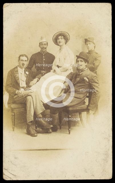 Amateur actors sitting down, surrounding a man in drag seated in the centre, [1910-1919?]. Creator: Unknown.