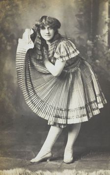An amateur actor in drag holds up his impressive folded dress to one side, (between 1910 and 1919?). Creator: Unknown