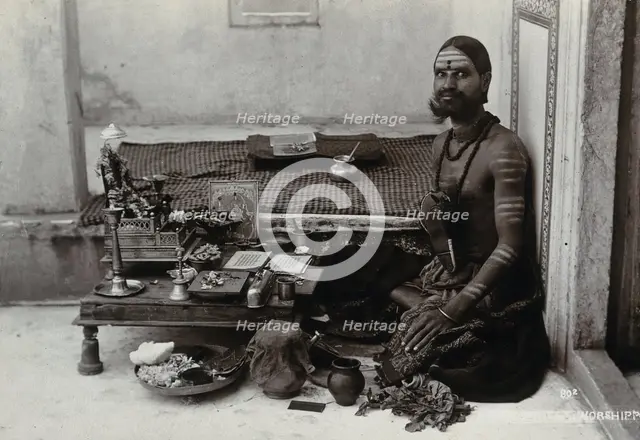 An Indian man wearing elaborate jewellery and make-up, squatting in front of a small table..., c1900 Creator: Unknown.