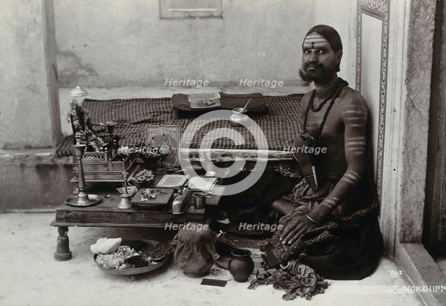 An Indian man wearing elaborate jewellery and make-up, squatting in front of a small table..., c1900 Creator: Unknown.