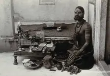 An Indian man wearing elaborate jewellery and make-up, squatting in front of a small table..., c1900 Creator: Unknown