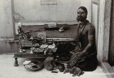 An Indian man wearing elaborate jewellery and make-up, squatting in front of a small table..., c1900 Creator: Unknown
