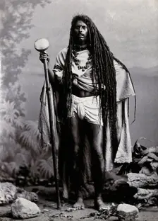 An Indian man with long hair, carrying a staff, in a studio setting, 1900. Creator: Unknown