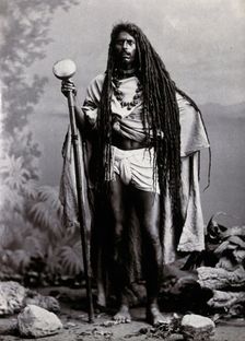 An Indian man with long hair, carrying a staff, in a studio setting, 1900. Creator: Unknown