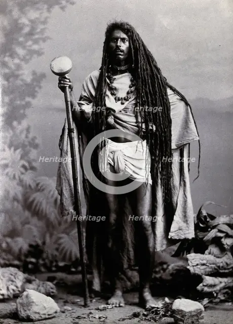 An Indian man with long hair, carrying a staff, in a studio setting, 1900. Creator: Unknown.