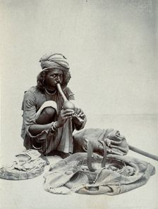 An Indian snake charmer squatting down playing his pipe, with two cobras in a basket in..., c1900. Creator: Unknown