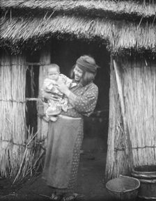 Ainu woman holding a child standing outside a hut, 1908. Creator: Arnold Genthe