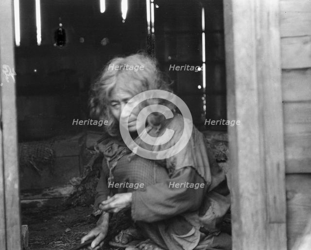Ainu woman seated in the doorway of a wooden hut, 1908. Creator: Arnold Genthe.