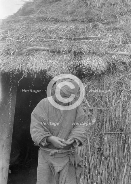 Ainu woman standing outside the entrance of a hut, 1908. Creator: Arnold Genthe.