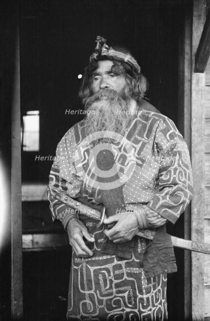 Ainu chief wearing a headdress and holding a sword standing in a doorway, 1908. Creator: Arnold Genthe.