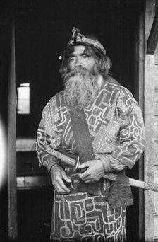Ainu chief wearing a headdress and holding a sword standing in a doorway, 1908. Creator: Arnold Genthe