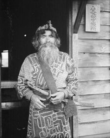 Ainu chief wearing a headdress and holding a sword standing in a doorway, 1908. Creator: Arnold Genthe