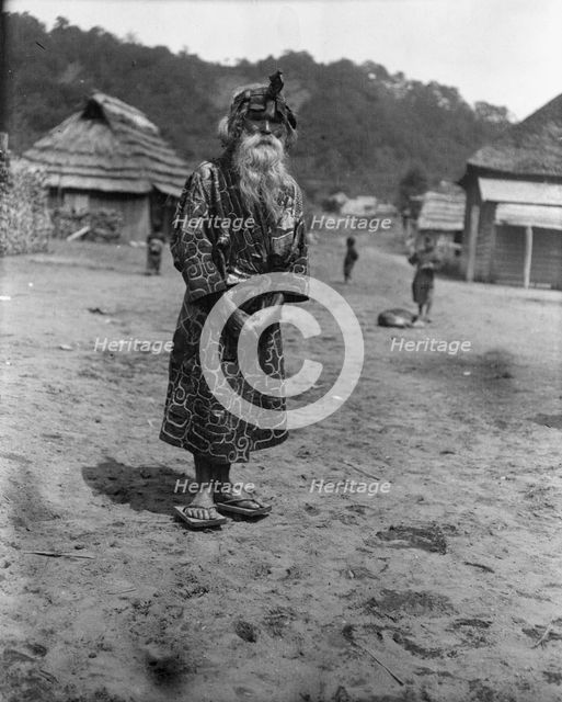 Ainu chief wearing a headdress standing in the village lane, 1908. Creator: Arnold Genthe.