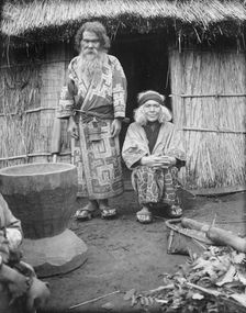 Ainu man and seated woman at the entrance of a hut, 1908. Creator: Arnold Genthe