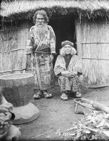 Ainu man and seated woman at the entrance of a hut, 1908. Creator: Arnold Genthe