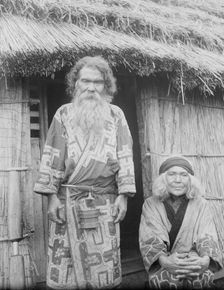 Ainu man and seated woman at the entrance of a hut, 1908. Creator: Arnold Genthe