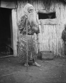 Ainu man holding a staff standing outside a hut, 1908. Creator: Arnold Genthe
