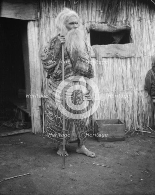 Ainu man holding a staff standing outside a hut, 1908. Creator: Arnold Genthe.