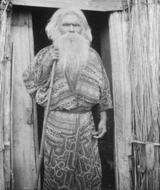 Ainu man holding a staff standing at the doorway of a hut, 1908. Creator: Arnold Genthe