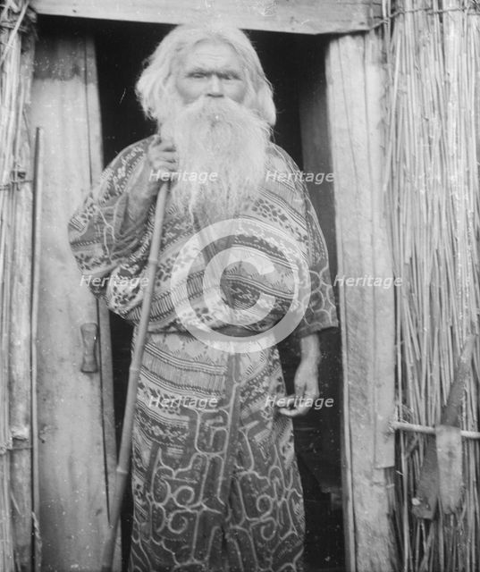 Ainu man holding a staff standing at the doorway of a hut, 1908. Creator: Arnold Genthe.
