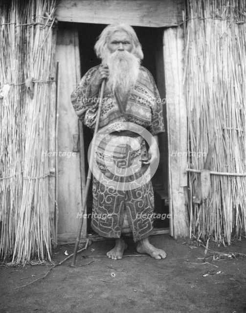 Ainu man holding a staff standing at the doorway of a hut, 1908. Creator: Arnold Genthe.