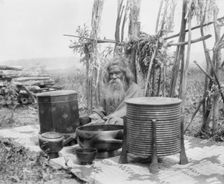 Ainu man seated outdoors on a mat covered with clay containers, 1908. Creator: Arnold Genthe