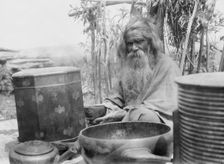 Ainu man seated outdoors on a mat covered with clay containers, 1908. Creator: Arnold Genthe
