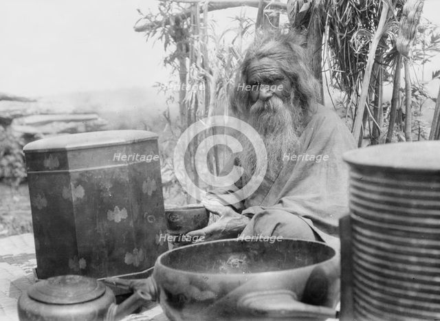 Ainu man seated outdoors on a mat covered with clay containers, 1908. Creator: Arnold Genthe.
