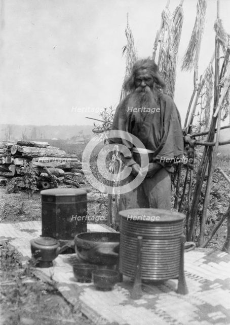 Ainu man standing outdoors on a mat covered with clay containers, 1908. Creator: Arnold Genthe.