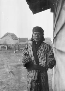 Ainu standing outside by the wall of a wooden hut, 1908. Creator: Arnold Genthe