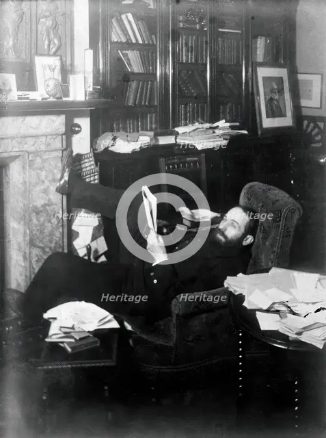 Arthur Schuster, lying on an armchair by a fireplace, with a photograph of Lord Kelvin..., c1901. Creator: Unknown.