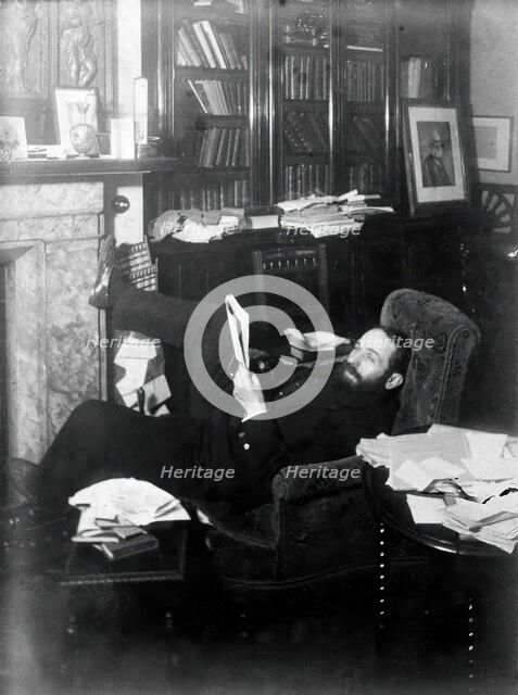 Arthur Schuster, lying on an armchair by a fireplace, with a photograph of Lord Kelvin..., c1901. Creator: Unknown.