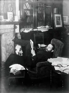 Arthur Schuster, lying on an armchair by a fireplace, with a photograph of Lord Kelvin..., c1901. Creator: Unknown