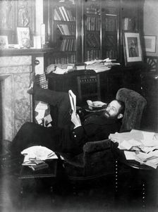 Arthur Schuster, lying on an armchair by a fireplace, with a photograph of Lord Kelvin..., c1901. Creator: Unknown