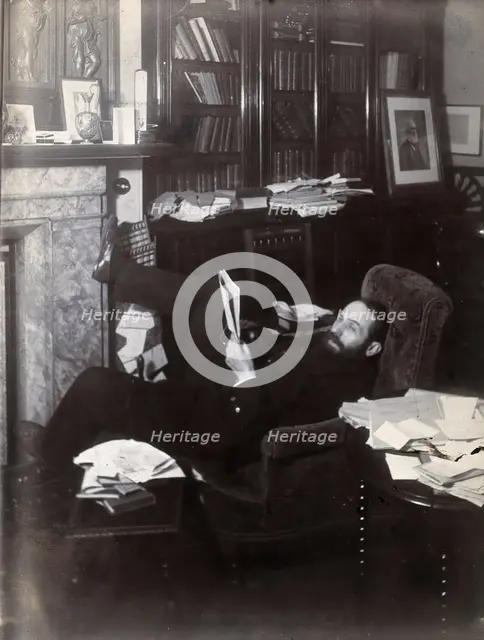 Arthur Schuster, lying on an armchair by a fireplace, with a photograph of Lord Kelvin..., c1901. Creator: Unknown.