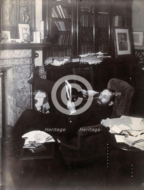 Arthur Schuster, lying on an armchair by a fireplace, with a photograph of Lord Kelvin..., c1901. Creator: Unknown.