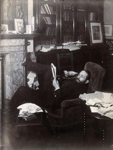 Arthur Schuster, lying on an armchair by a fireplace, with a photograph of Lord Kelvin..., c1901. Creator: Unknown