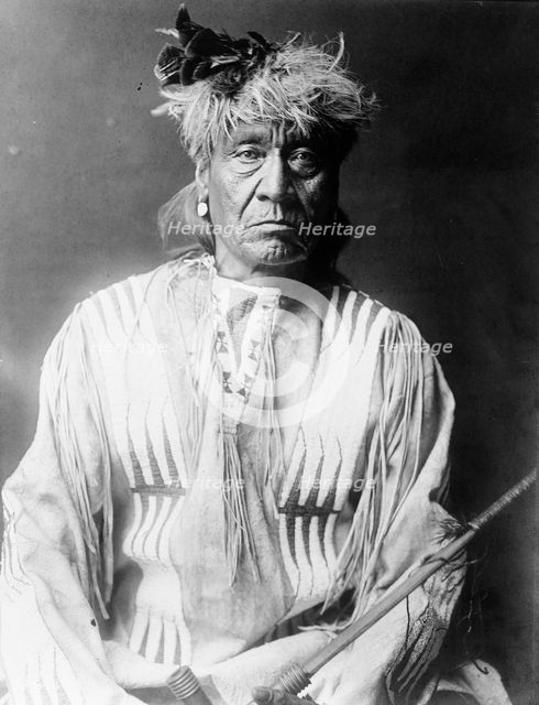 Atsina Indian, Red Whip, half-length portrait, seated, facing front, wearing feather..., c1908. Creator: Edward Sheriff Curtis.