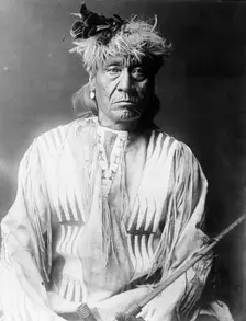 Atsina Indian, Red Whip, half-length portrait, seated, facing front, wearing feather..., c1908. Creator: Edward Sheriff Curtis