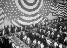 Banquet Scene with Flag Draping And Men Seated Holding Up Listening Device To One Ear, c1913-1917. Creator: Harris & Ewing