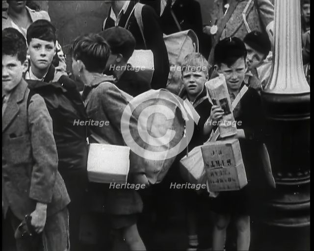 British Child Evacuees Standing on a Pavement With Bags and Boxes as Adults are Standing..., 1939. Creator: British Pathe Ltd.