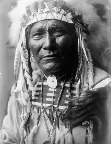 Ghost Bear, Crow Indian, Montana, head-and-shoulders portrait, facing front, feather..., c1908. Creator: Edward Sheriff Curtis