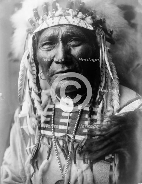 Ghost Bear, Crow Indian, Montana, head-and-shoulders portrait, facing front, feather..., c1908. Creator: Edward Sheriff Curtis.