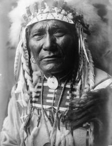 Ghost Bear, Crow Indian, Montana, head-and-shoulders portrait, facing front, feather..., c1908. Creator: Edward Sheriff Curtis