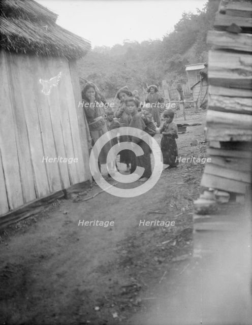 Group of Ainu children standing in a passageway between huts, 1908. Creator: Arnold Genthe.