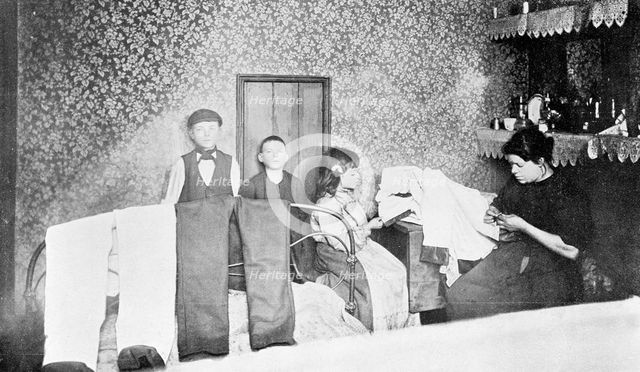 Domestic scene showing a mother with her children finishing trousers, London, c1900. Creator: Unknown.