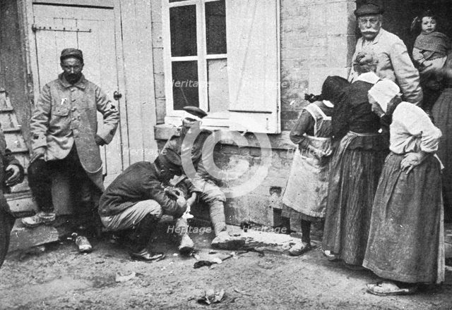 'Derriere L'ennemi refoule; Un major soignant un allemand blesse que des villageois..., 1915 (1924). Creator: Unknown.